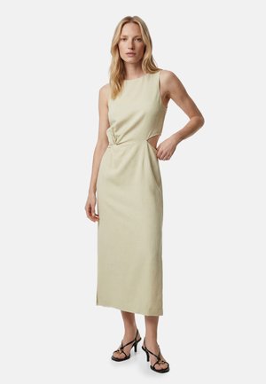 Kvinde iført ærmeløs beige midi-kjole med sidesnit og sorte højhælede sandaler, stående foran en ensfarvet hvid baggrund.