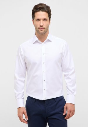 Mand iført en hvid, knaplukket, langærmet skjorte og mørkeblå bukser, stående foran en ensfarvet lys baggrund.