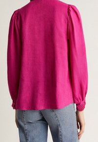 Blusa fucsia con maniche lunghe, spalle arricciate e bordi in pizzo ai polsini. Indossata con jeans azzurri, che mettono in mostra un tessuto testurizzato.