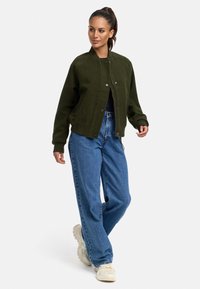 Olivgrön bomberjacka med dragkedja framtill, ribbade ärmslut och oversized passform, i kombination med lösa blå denimjeans och beige sneakers.
