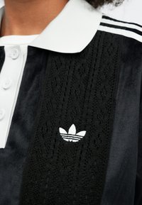 Tricou polo alb și negru, cu un panou texturat negru, nasturi albi și un logo Adidas brodat alb pe piept.