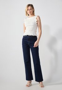 Hvit smocket topp med volangermar er parret med mørkeblå vide jeans og lyse beige sandaler med høy hæl. Enkel, strukturert design.