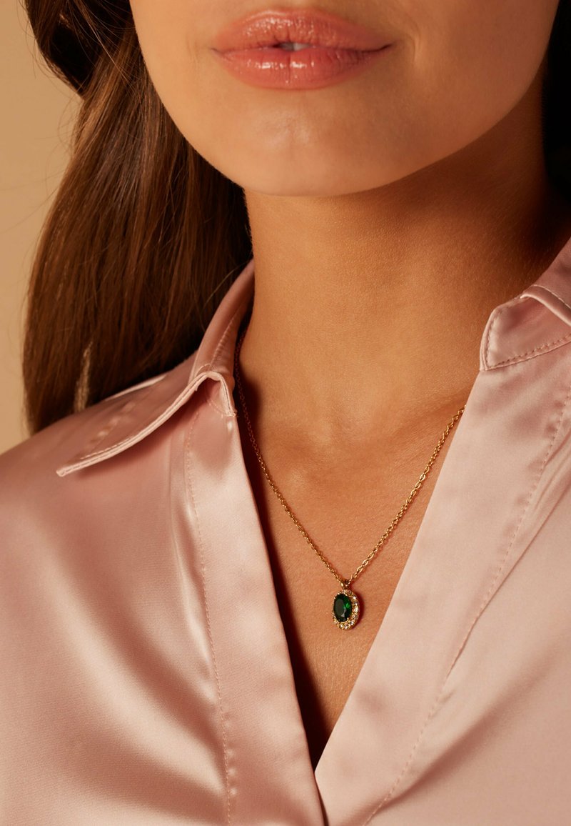Gouden ketting met een ovale groene hanger omringd door een cirkelvormige rhinestone-accent, gedragen over een roze satijnen shirt.