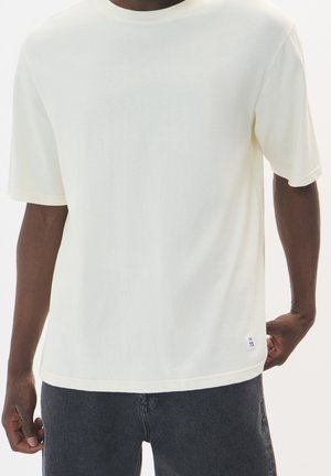 Man draagt een effen off-white T-shirt met korte mouwen en donkere spijkerbroek, staand met één hand achter zijn rug.