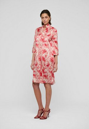 Nainen seisoo harmaata taustaa vasten yllään polvipituinen vaaleanpunainen satiininen kukkamekko, jossa on 3/4-hihat, ja punaiset korkokengät.