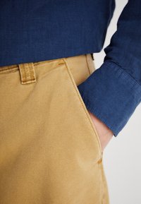 Μπλε λινό πουκάμισο με μακριά μανίκια, συνδυασμένο με καπέλ μπεζ βαμβακερού παντελονιού που διαθέτει πλαϊκή τσέπη και υφασμάτινη υφή.