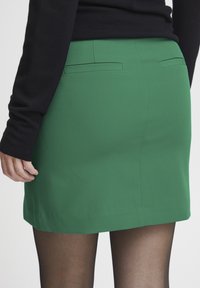 Fustă verde cu o textură netedă, având două buzunare în spate și un croi drept. Poartă colanți negri, evidențiind detaliile tivului.