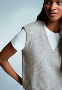 Beige gestrickte Weste mit V-Ausschnitt und gerippten Kanten, getragen über einem weißen kurzärmeligen Hemd. Weicher Stoff, lässige Passform.