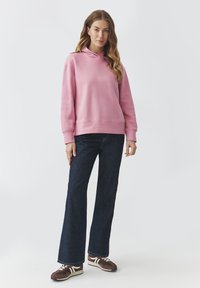 Felpa rosa con cappuccio e cordoncino, abbinata a jeans a zampa d'elefante in denim scuro e sneakers marroni con dettagli bianchi.