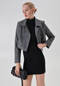 Donna che indossa un giubbotto corto grigio a doppio petto sopra un vestito nero a collo alto, mentre tiene in mano una borsa nera con borchie su uno sfondo neutro.