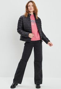 Zwarte gewatteerde jas met ritssluiting, gecombineerd met een roze sweatshirt met witte tekst, en zwarte wijde jeans. Zwarte platformlaarzen.