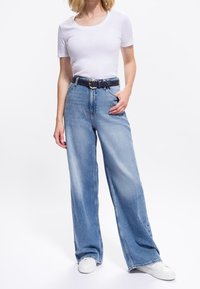 Femeie purtând tricou alb cu mâneci scurte, blugi albaștri cu talie înaltă și croială largă, cu o curea împletită închisă la culoare, și adidași albi, stând cu mâna în buzunar.