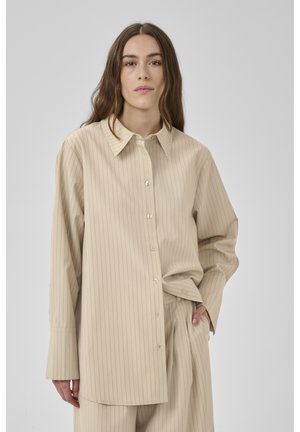 Ung kvinde med langt brunt hår iført en beige oversized skjorte med nålestriber og matchende bukser foran en ensfarvet baggrund.