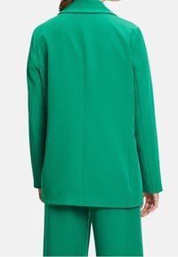 Persona vistiendo un blazer de manga larga verde brillante y pantalones a juego, vista desde atrás sobre un fondo claro.