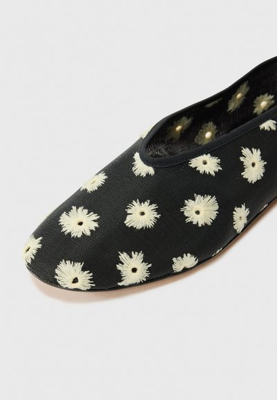 Zapato de lona negro con un patrón floral cremoso en toda la superficie. Puntera redondeada y suela flexible, con una textura suave y diseño sin cordones.