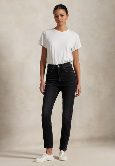 Jonge vrouw staande, draagt een wit shirt met korte mouwen, zwarte high-waist jeans en witte sneakers tegen een effen achtergrond.