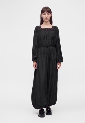 Femme aux cheveux bruns et raides portant une longue robe noire avec des détails en dentelle et des manches bouffantes, associée à des chaussures noires, debout sur un fond blanc.
