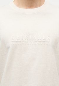 Κοντινό πλάνο σε λευκό πουκάμισο με στρογγυλή λαιμόκοψη, με ανάγλυφο κείμενο: "NINETEEN NINETY ORIGINALS 1990 BROUGHT TO YOU BY JACK & JONES."