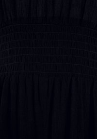 Svart klänning med en smockad midja, som har ett texturerat tyg och en flödande kjol. Materialet verkar mjukt och lätt.