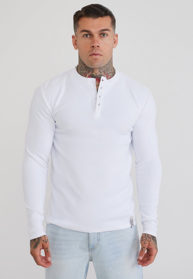 Man with neck and hand tattoos wears long-sleeve white henley shirt and light blue jeans, standing against plain light background.
