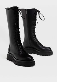 Un par de botas altas de cuero negro con cordones, suelas gruesas y cremalleras laterales sobre un fondo blanco.