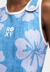 Κοντινή λήψη μπλε φλοράλ αμάνικου τοπ με λευκή λεπτομέρεια και λογότυπο Roxy στο στήθος, φορεμένο από άτομο με μέτρια σκούρο τόνο δέρματος.