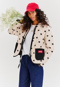 Prešívaná krémová bunda s čiernym vzorom srdca, čiernymi vreckami, sťahovacou šnúrkou, kombinovaná s modrými džínovými nohavicami a červenou čiapkou.