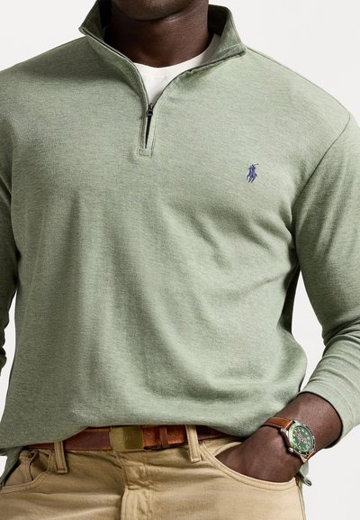 Green, long-sleeve pullover with a zip collar, featuring a chest logo. Paired with a brown belt and a green-faced watch.