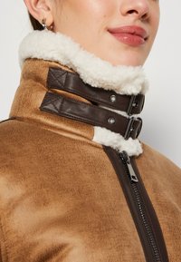 Giubbotto in finta pelle marrone con colletto in shearling color crema, chiusure a doppia fibbia e zip centrale.