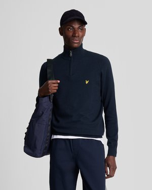 Navy zip-up sweater with a high neck, featuring a small yellow logo. Paired with a dark cap and carrying a navy shoulder bag.