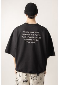 Czarna, duża koszulka z krótkim rękawem, z białym napisem na plecach. Materiał wygląda na miękki, o luźnym kroju i swobodnym designie.