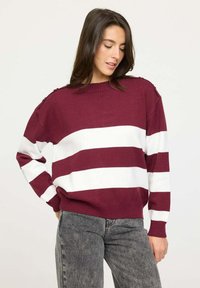 Pull maroon et blanc à rayures en tricot doux. Présente un col rond et des boutons à l'épaule pour un détail supplémentaire. Associé à un jean gris.
