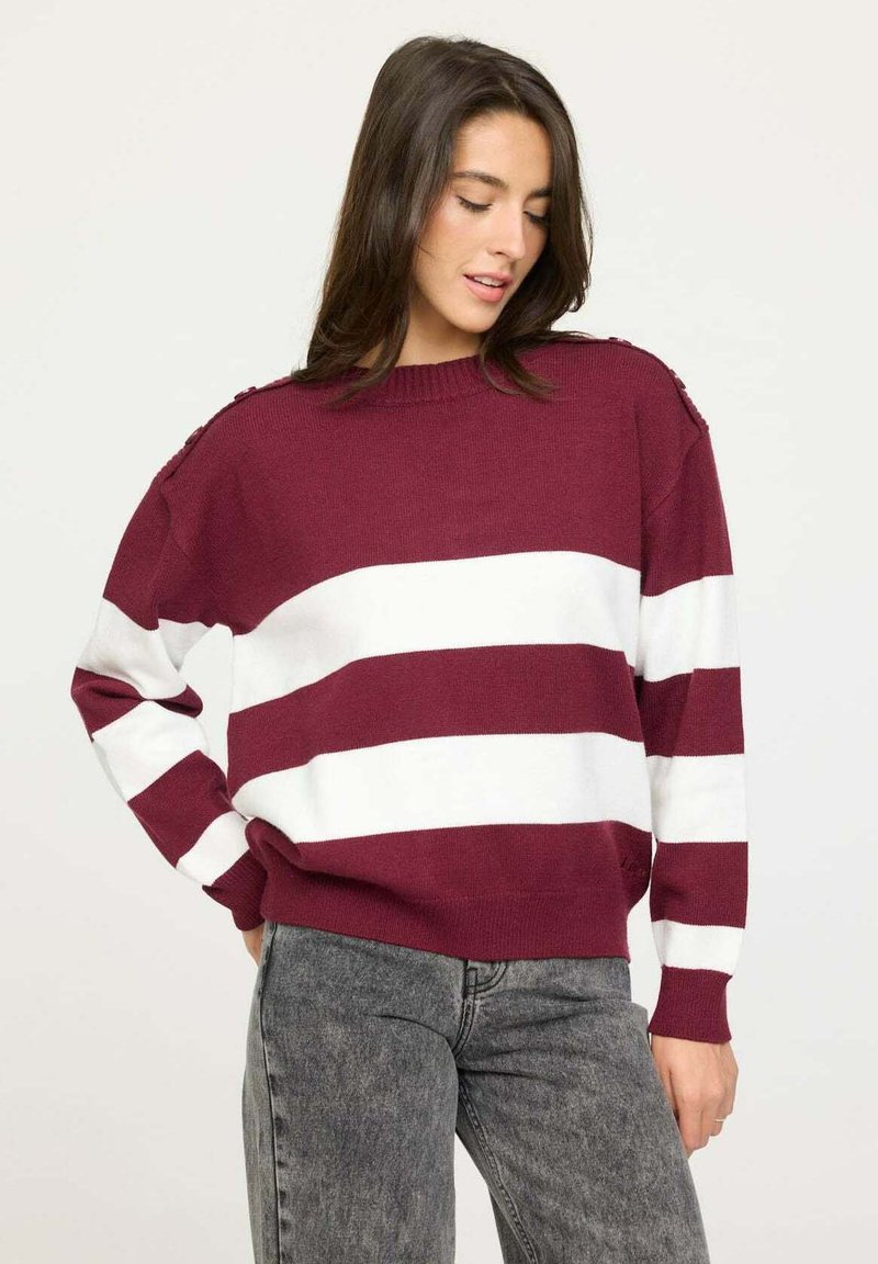 Pull maroon et blanc à rayures en tricot doux. Présente un col rond et des boutons à l'épaule pour un détail supplémentaire. Associé à un jean gris.