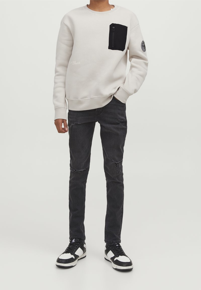 Personne portant un sweat-shirt beige clair avec une poche zippée noire, un jean slim noir usé et des baskets noir et blanc, debout.