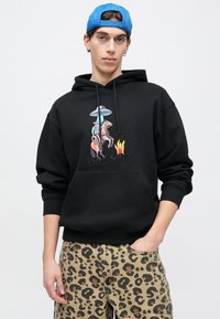 Μαύρο φούτερ με κουκούλα με γραφικό ενός καουμπόι πάνω σε άλογο κάτω από ένα UFO και φλόγες. Περιλαμβάνει τσέπη καγκουρό και κορδόνια στη κουκούλα.