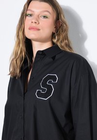 Camicia nera con bottoni in cotone, con colletto, dotata di una lettera "S" ricamata in bianco sul lato sinistro, texture liscia.