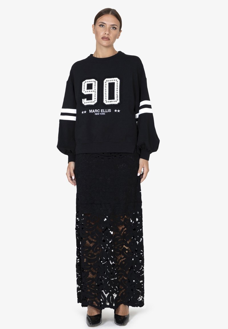 Sweat-shirt noir à coupe oversize, manches rayées blanches et numéro '90' en strass. Associé à une longue jupe en dentelle noire et des chaussures brillantes.