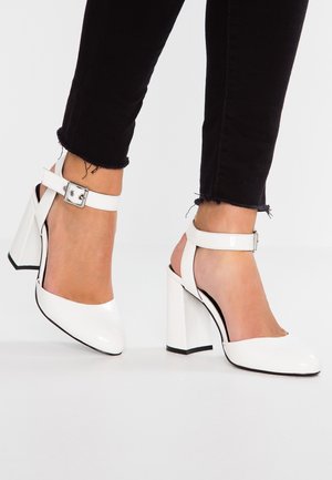 Hvide højhælede sandaler i laklæder med ankelrem, firkantet blokhæl og kontrasterende sorte detaljer langs sålen.