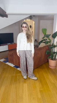 Baltas mygtukais užsegamas kardiganas, pilkos raštuotos platėjančios kelnės ir šviesios spalvos batai, stovint kambaryje su ruda sofa ir augalais.