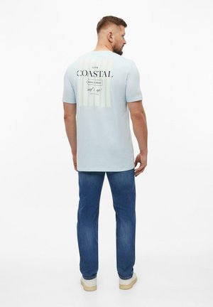 Mann trägt hellblaues T-Shirt mit dem Schriftzug „COASTAL“ und blaue Jeans, steht mit dem Rücken zur Kamera vor weißem Hintergrund.