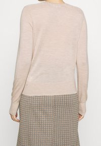 Pull à manches longues beige clair avec une texture douce. Il présente un col rond et des poignets ajustés, associé à une jupe à carreaux dans des tons terreux.