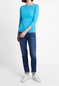 Kvinna som bär en ljusblå långärmad topp, mörkblå uppvikta jeans och vita sneakers, stående mot en vit bakgrund.