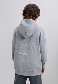Grijze hoodie met een zachte textuur, met een geborduurd schedel- en kruisbeenderenontwerp op de achterkant en "Scalpers" in witte letters eronder.