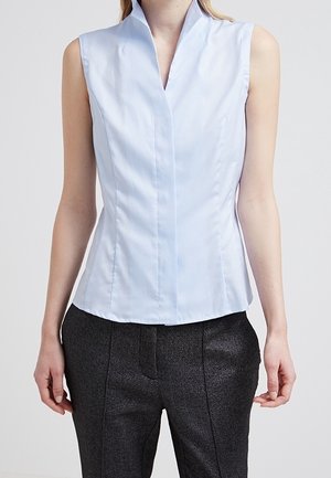 Bez rękawów, dopasowana, jasnoniebieska bluzka z wysokim kołnierzem, połączona z ciemnymi, teksturowanymi, dopasowanymi spodniami na stojącej osobie.