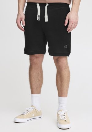 Shorts en polaire noirs avec une taille élastique à cordon de serrage, des poches latérales et un logo sur la jambe gauche. Associés à des baskets beiges et des chaussettes blanches.