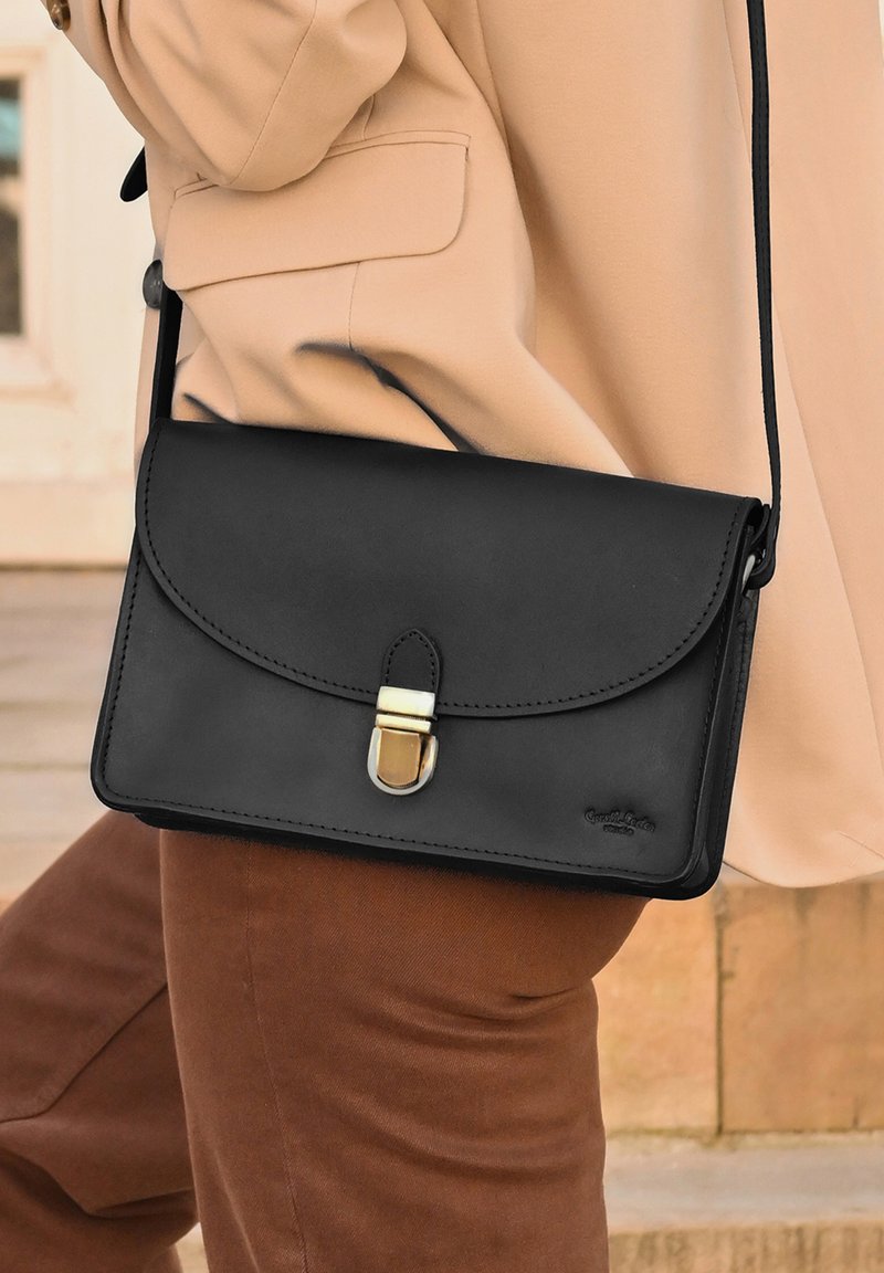 Črna usnjena torbica čez ramo z zaponko v zlati barvi in preklopnim pokrovom. Gladek tekstura, zaobljeni robovi in vtisnjen logotip blagovne znamke na sprednji strani.