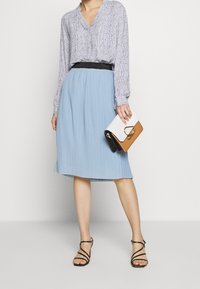 Donna con una blusa a maniche lunghe con motivo, infilata in una gonna plissettata di colore azzurro chiaro, che tiene una pochette bicolore e indossa tacchi neri con cinturini.