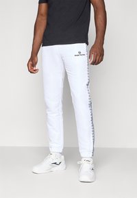 Mand i hvide Sergio Tacchini træningsbukser med logo-tape på siderne, sort t-shirt og hvide sportssko står på en ensfarvet baggrund.