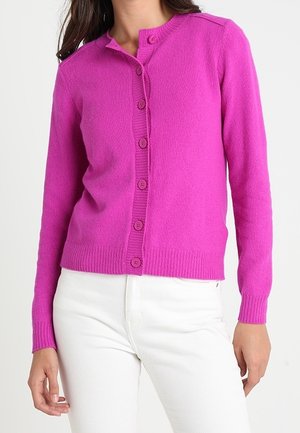 Vrouw draagt een felroze gebreid vest met knopen en lange mouwen, gecombineerd met een witte broek, tegen een effen achtergrond.