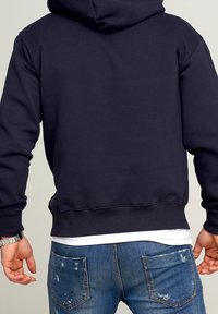 Sudadera azul marino con un bolsillo canguro, hecha de tela suave. Cuenta con una capucha forrada y puños acanalados. Llevada sobre unos jeans azules desgastados.
